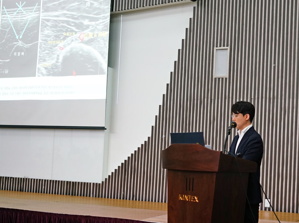킨텍스 로고가 있는 강단에서 한 남성이 발표하고 있으며, 뒤 스크린에는 초음파 영상과 의료 관련 자료가 표시되어 있다 — 대한한의영상학회 초음파 약침·도침 강의 | 서울 바로한의원 안태석 원장
