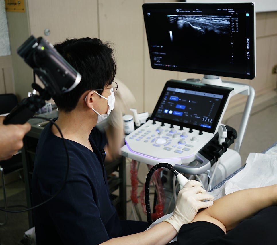 마스크를 착용한 의료진이 환자의 팔에 초음파 탐촉자를 대고 초음파 검사를 진행하는 모습 — 초음파 유도하 약침 시술 | 서울 바로한의원