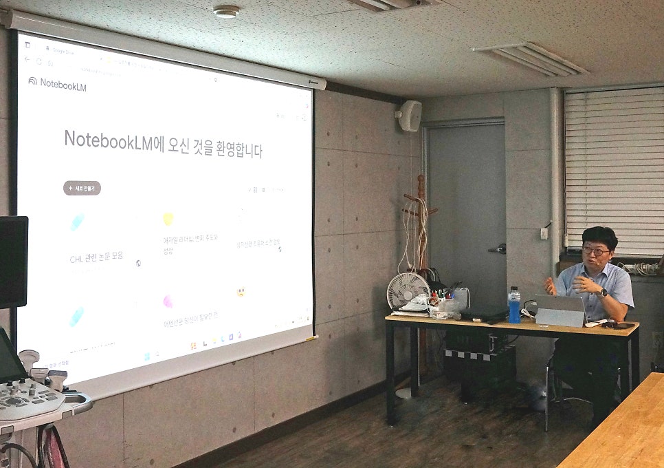 강사가 노트북LM 화면을 띄운 프로젝터 앞에서 발표하고 있으며, 왼쪽에 초음파 기기가 놓여 있다 — 대한한의영상학회 초음파 약침·도침 강의 | 서울 바로한의원
