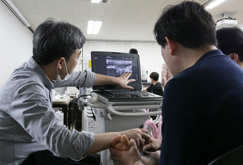 마스크를 쓴 사람이 초음파 기기 화면의 초음파 영상을 가리키고 있고, 다른 사람의 발에 초음파 탐촉자가 놓여 있다 — 초음파 유도하 약침 시술 | 서울 바로한의원