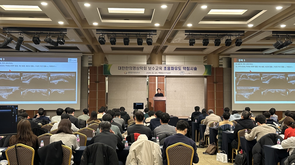 강당에서 강사가 연단에 서서 청중에게 강연하고 있으며, 양쪽 스크린에는 초음파 영상이 보인다 — 대한한의영상학회 초음파 약침·도침 강의 | 서울 바로한의원