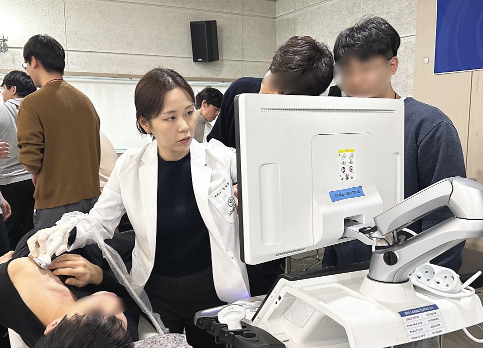 한의사가 환자의 상체에 초음파 기기를 대고 모니터를 보며 진료하고 있으며, 주변 사람들이 이를 지켜보고 있습니다 — 초음파 유도하 약침 시술 | 서울 바로한의원