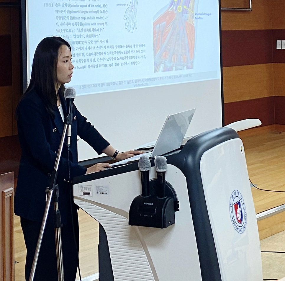 한 여성이 강단에서 노트북과 마이크 앞에서 발표하고 있으며, 뒤 스크린에는 해부학 그림과 글이 보인다 — 대한한의영상학회 초음파 약침·도침 강의 | 서울 바로한의원 문지현 원장