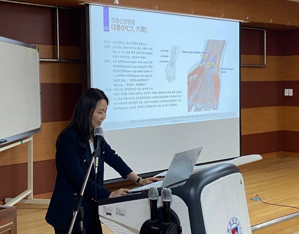 한 여성이 강단에서 노트북과 마이크가 있는 연단에 서서 발표하고 있으며, 뒤편 스크린에는 손목 해부학 다이어그램과 정중신경병증 관련 한글 텍스트가 보인다 — 대한한의영상학회 초음파 약침·도침 강의 | 서울 바로한의원 문지현 원장