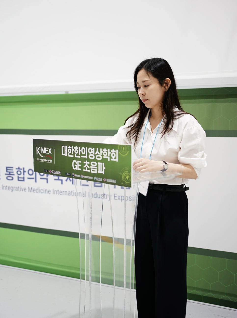 여성이 투명 연단에 서서 아래를 보고 있으며, 연단에는 '대한한의영상학회 GE 초음파'라고 적힌 표지가 놓여 있다 — 대한한의영상학회 초음파 약침·도침 강의 | 서울 바로한의원 문지현 원장