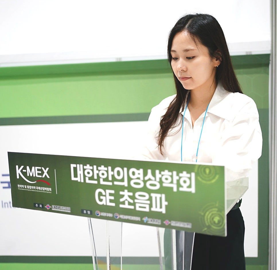 흰 셔츠를 입은 여성이 K-MEX 대한한의영상의학회 GE 초음파라고 적힌 연단 뒤에 서 있다 — 대한한의영상학회 초음파 약침·도침 강의 | 서울 바로한의원 문지현 원장