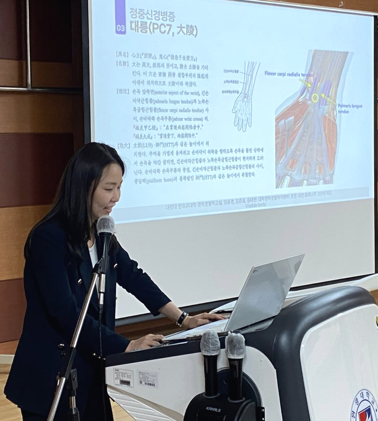 한 여성이 강단에서 발표하며, 뒤쪽 스크린에 손목 해부학 다이어그램과 한의학 관련 내용이 보인다 — 대한한의영상학회 초음파 약침·도침 강의 | 서울 바로한의원 문지현 원장