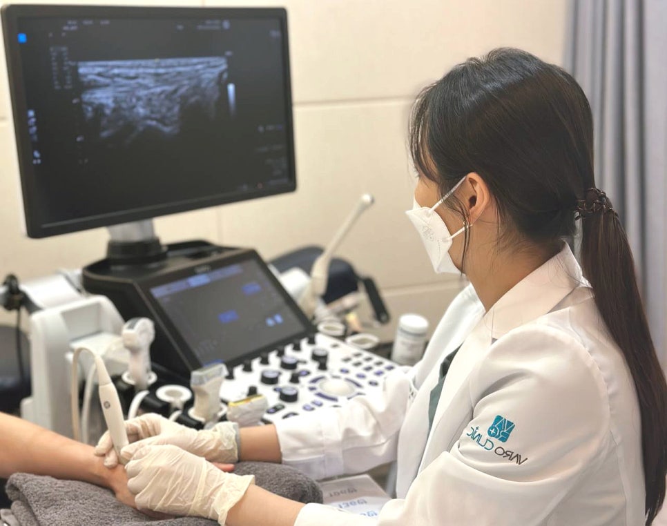 마스크를 쓴 의료진이 환자의 손목에 초음파 기기를 대고 검사하며, 모니터에 초음파 영상이 보인다 — 초음파 정밀 진단 | 서울 바로한의원 초음파 약침·도침·침도
